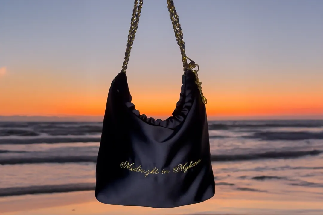 Sac de soirée en satin bleu nuit brodé « Midnight in Mykonos » au fil d'or, devant un coucher de soleil